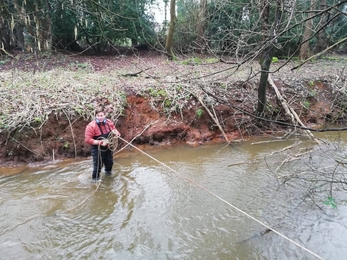 Joshua stands in a river holding a rope that is attached to the bank