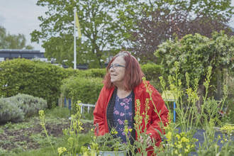 A woman stands by a vegetable patch in a community garden, surrounded by greenery, holding a bowl of freshly collected vegetables.