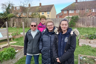 Four people stand in a community garden with raised beds to either side. 