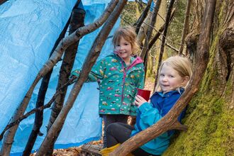 Two girls in a den made of sticks leaning against a tree