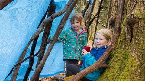 Two girls in a den made of sticks leaning against a tree