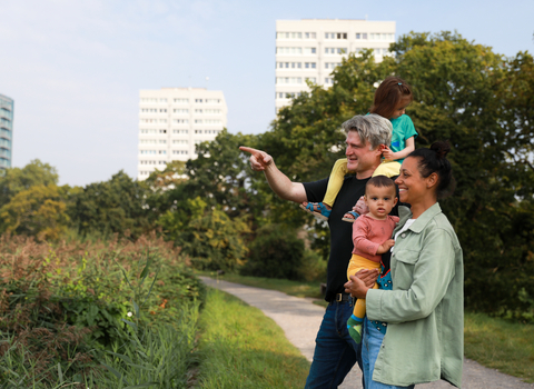 A family of four are at a nature reserve with buildings visible in the background. The father is pointing to show his partner and children something in the distance