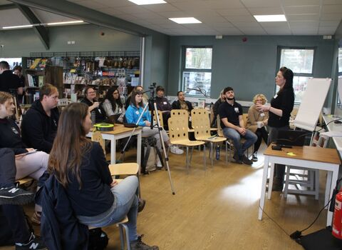a group of people gathered indoors watching a speaker present. 