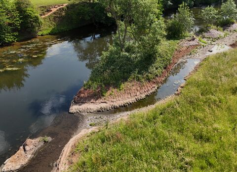 Aerial view of a river bend bordered by grassy banks and trees, with a narrow gravel path following the shoreline. The water curves around a small vegetated peninsula